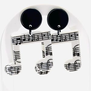 May include: White acrylic earrings shaped like music notes with a black and white sheet music design. The earrings have black round dangles.
