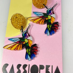 May include: A pair of colourful hummingbird earrings with gold glitter studs. The hummingbirds feature a stained-glass design with vibrant hues of blue, yellow, green, and red. The earrings are displayed on a pink card with the text "CASSIOPEIA AND CO."