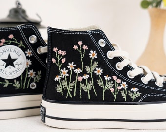 Puede incluir: Zapatillas Converse negras de caña alta con costuras blancas y un logotipo de estrella blanca. Las zapatillas están bordadas con flores blancas y rosas.