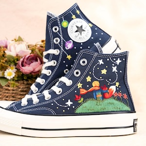 May include: Navy blue high-top sneakers with white laces and soles. The sneakers feature embroidered designs of stars, planets, and a scene from The Little Prince. A floral arrangement sits in the background.