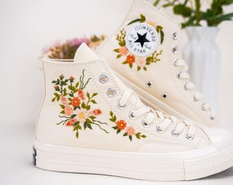 May include: A pair of off-white high-top trainers with floral embroidery. The trainers have a white rubber sole and a black Converse All Star logo on the side.