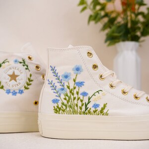 Puede incluir: Zapatillas Converse blancas de caña alta con suela de plataforma. Las zapatillas están bordadas con flores azules y hojas verdes. El logotipo de Converse All Star está bordado en el lateral de la zapatilla.