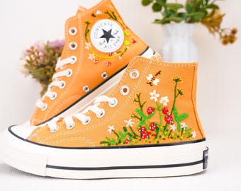 Puede incluir: Un par de zapatillas de lona de caña alta con diseños florales bordados. Las zapatillas son de color naranja con suelas y cordones blancos. La zapatilla izquierda tiene el logotipo de Converse en el lateral.