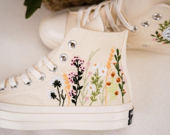 May include: A pair of off-white high-top canvas sneakers with embroidered flowers in shades of pink, green, and yellow. The sneakers have white laces and a white rubber sole.