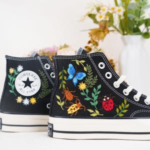 Könnte beinhalten: Schwarze Converse-Sneaker mit hohem Schaft, bestickt mit Blumen, Blättern und Insekten. Das Converse All Star Logo ist an der Seite des Schuhs sichtbar.