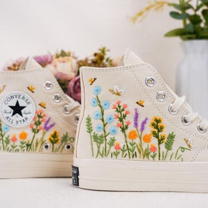 May include: A pair of off-white high-top canvas sneakers featuring colorful floral embroidery. The design includes various wildflowers in shades of blue, orange, pink, and green, along with small embroidered bees and butterflies on the side panels.