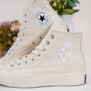 Puede incluir: Un par de zapatillas Converse de caña alta de color blanco roto con bordados florales blancos y un logotipo Converse All Star negro.
