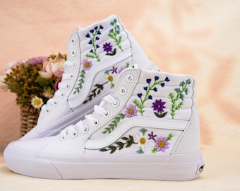 May include: Pair of white high-top sneakers with embroidered floral designs in purple, blue, and yellow. The sneakers have white laces and soles. A basket of flowers is in the background.