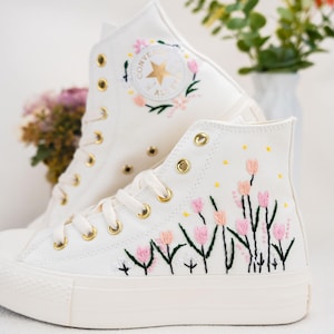 Könnte beinhalten: Weiße Converse-Sneaker mit hohem Schaft und gestickten rosa und gelben Blumen. Die Sneaker haben goldene Ösen und eine weiße Gummisohle.