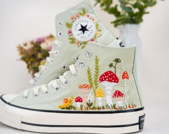 May include: Sage green high-top Converse sneakers with embroidered mushrooms, flowers, and leaves. The Converse logo is visible on the side of the shoe.