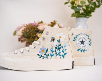Peut inclure: Une paire de baskets Converse montantes de couleur blanc cassé avec des broderies florales. La broderie présente des fleurs et des feuilles bleues et vertes. Les baskets ont une semelle en caoutchouc blanche et un logo Converse All Star noir sur le côté.