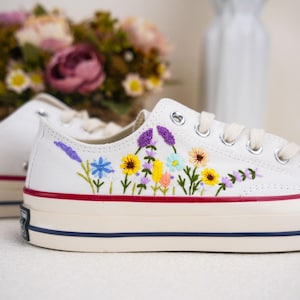 May include: White canvas sneakers with embroidered flowers in shades of purple, blue, yellow, and pink. The flowers are arranged in a row across the shoe.