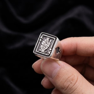 May include: A silver ring with a King of Spades card design. The ring has a rectangular face with a detailed engraving of a skeleton king holding a sword. The spade symbol is cut out on the side of the ring.