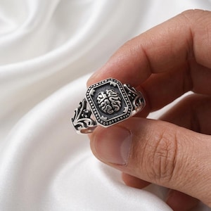 May include: A silver ring featuring a detailed brain design within a rectangular frame. The ring has an ornate, vintage-inspired style with decorative flourishes on the band. The ring is held up by a hand.