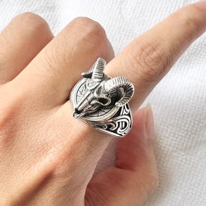 Może przedstawiać: Srebrny pierścień z motywem czaszki barana. Pierścień ma szczegółowy, teksturowany wygląd z zakrzywionymi rogami i okrągłym emblematem. Obręcz pierścienia ma ozdobny, wzorzysty design.