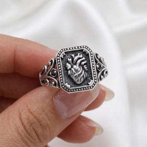 Puede incluir: Un anillo de plata con un diseño detallado de corazón anatómico dentro de un marco rectangular. El anillo tiene una banda ornamental de filigrana con elementos decorativos. El corazón es el punto focal, representado en un estilo realista. El anillo es sostenido por una mano.