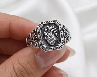 May include: A silver ring featuring a detailed anatomical heart design within a rectangular frame. The ring has an ornate, filigree band with decorative elements. The heart is the focal point, rendered in a realistic style, and the ring is held by a hand.