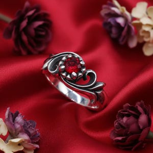 May include: A silver ring featuring a round, faceted red gemstone set in a floral-inspired mounting with intricate, swirling metalwork details. The band has a polished finish and is accented by dark, recessed areas that highlight the scrollwork.