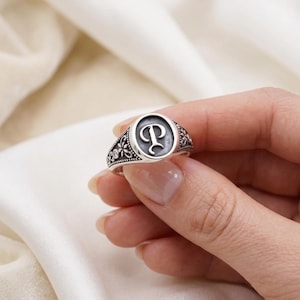 May include: A silver signet ring with the letter "P" in a circular design. The ring has floral and dot detailing around the edges and is held by a hand.