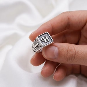 Könnte beinhalten: Ein silberfarbener Siegelring mit quadratischer Front, auf der der Buchstabe H in einer gotischen Schrift eingraviert ist. Die Seiten des Rings sind mit komplexen, dunkel oxidierten floralen Rankenmustern verziert, die sich vom polierten Metall abheben.
