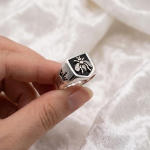 Op de afbeelding: Een zilveren zegelring met een zwart geëmailleerd schild met een gedetailleerd bijenontwerp. De ring heeft een kroondetail aan de zijkant. De ring wordt in een hand gehouden.