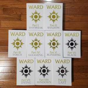 May include: Nine books with white covers are arranged on a wooden surface. Each book has the title "WARD" in gold lettering, along with a sunburst design and a part number and title. The titles include "DAYBREAK," "SHADOW," "TORCH," and "BLACK."