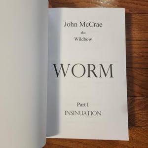 Puede incluir: Libro abierto con el título "WORM" en letras negras grandes. El nombre del autor, John McCrae, y el alias "Wildbow" están encima del título. El texto "Part I INSINUATION" está debajo.