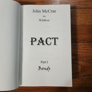 May include: Open book with the title page of "PACT" by John McCrae, also known as Wildbow. The text is in black font on a white page. The book is open on a wooden surface.
