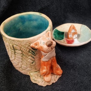 May include: A ceramic mug with a brown dog figurine attached to the side. The mug has a green and brown textured design. The mug comes with a matching saucer with a small house figurine in the center.