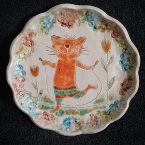 May include: A white ceramic plate with a scalloped edge features a hand-painted illustration of an orange cat jumping rope. The cat is wearing a green and orange striped swimsuit. The plate is decorated with a colorful floral border.