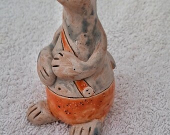 May include: A ceramic figurine of a gray cat with orange shorts and green eyes. The cat is standing on its hind legs and has its arms crossed.