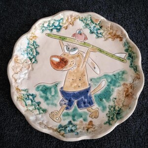 May include: A ceramic plate with a cartoon dog wearing blue shorts and holding a surfboard with a bird on top. The plate has a white background with green, brown, and blue accents.