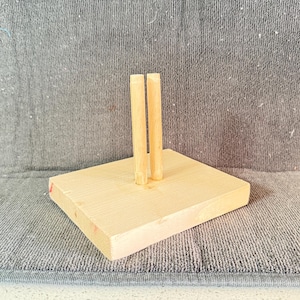May include: A small, unfinished wooden display stand with two vertical dowels. The stand is rectangular and made of light-colored wood, with a natural grain. The dowels are also made of wood and are positioned in the center of the stand.