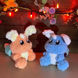 May include: Two handmade crochet plush toys. One is orange with cream-colored ears and a rainbow tail, the other is blue with white ears and a rainbow tail. Both have black eyes and are sitting on a surface.
