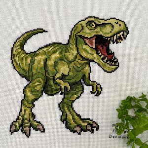 Puede incluir: Punto de cruz de un Tyrannosaurus Rex verde con la boca abierta, revelando detalles rojos y negros. El dinosaurio está sobre un fondo blanco. El bordado está detallado con varios tonos de verde.