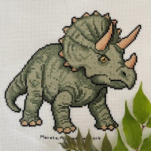 Pode incluir: Ponto cruz de um dinossauro Triceratops em tons de verde, bege e preto. O dinossauro tem três chifres e uma gola, sobre um fundo branco. O texto "Merete; All aboard the ark" é visível na parte inferior.