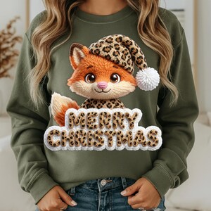 May include: An olive green sweatshirt featuring a knitted fox wearing a leopard print hat with a white pom-pom. The fox is holding a sign that says "MERRY CHRISTMAS" in a leopard print design. The sweatshirt is a festive holiday item.