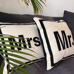 Könnte beinhalten: Zwei quadratische Dekokissen mit cremefarbener Webstruktur und einem dicken schwarzen Rand. Ein Kissen zeigt den schwarzen gestickten Text "Mrs." und das andere den schwarzen gestickten Text "Mr." in einer fetten, serifenlosen Schriftart.