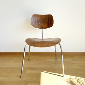Könnte beinhalten: Ein Stuhl mit einer geschwungenen Sitzfläche aus mittelbraunem Holz und einer passenden Rückenlehne. Die Rückenlehne ist durch eine einzelne vertikale Metallstütze mit dem Sitz verbunden. Der Stuhl hat vier schlanke, silberfarbene Metallbeine.