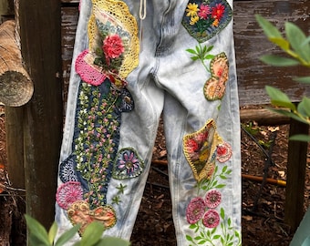 May include: Light-wash denim jeans adorned with embroidered patches. The patches showcase floral designs, hearts, and other decorative elements. The jeans have a drawstring waist and frayed hems, offering a unique, artistic aesthetic.