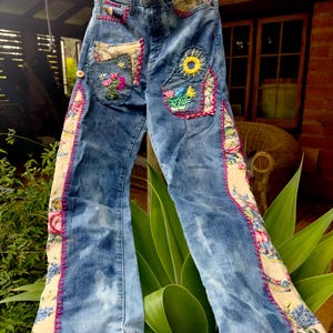 May include: Blue denim jeans with colorful embroidered patches and floral fabric accents. The jeans feature a sun, flowers, and other decorative elements. The seams are accented with pink stitching.