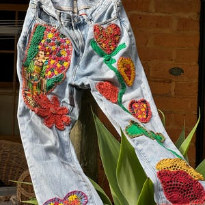 May include: Light blue denim jeans with colorful crochet floral and heart appliqués. The jeans feature a detailed crochet design of a woman, flowers, and hearts, with a crochet trim at the hem.