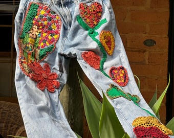 May include: Light blue denim jeans adorned with vibrant crochet floral and heart appliqués. The jeans showcase a detailed crochet design of a woman, flowers, and hearts, with a crochet trim at the hem.
