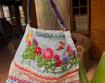 May include: A light blue denim shoulder bag featuring colorful floral embroidery, a small butterfly motif, and a sun icon. The lower section is decorated with layers of beige lace, green and white gingham fabric, and black and white checkered trim.