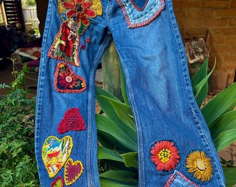 May include: Blue denim jeans decorated with embroidered patches in various shapes and colours. The patches include hearts and flowers. The jeans have a relaxed fit and are embellished with red stitching.