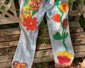 May include: Light blue denim jeans with colorful crochet embellishments. Features include a large flower, hearts, and a figure on the left leg. The jeans are decorated with red, yellow, green, and orange crochet details.