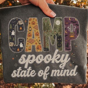 Puede incluir: Una camiseta gris oscuro con la palabra CAMP en letras grandes rellenas de patrones de fantasmas, calabazas, bosques y brujas. Debajo, el texto spooky state of mind está impreso en una fuente cursiva blanca con un contorno sutil.