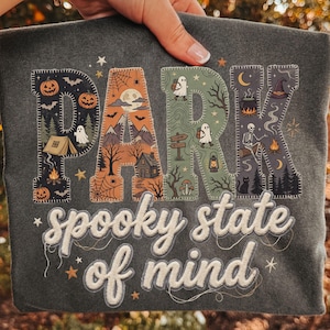 Puede incluir: Una camiseta gris oscuro con la palabra PARK en letras grandes rellenas de ilustraciones de Halloween y acampada, incluyendo calabazas, fantasmas, montañas y un esqueleto. Debajo, el texto spooky state of mind está impreso en blanco.
