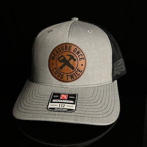 May include: A grey and black trucker hat featuring a circular brown leather patch on the front. The patch displays a graphic of a crossed hammer and saw with the text "MEASURE ONCE CUSS TWICE". A black sticker on the brim reads "RICHARDSON 112".