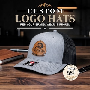 May include: A grey and black trucker hat featuring a brown leather-like patch on the front with the text "YOUR LOGO HERE". The hat has a structured crown, a curved brim with a black sticker, and a mesh back. A circular badge indicates youth sizes.